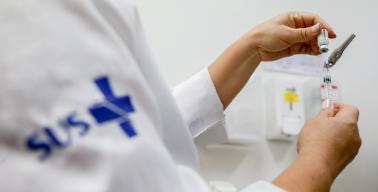 imagem de uma pessoa com jaleco do SUS, pegando uma vacina para aplicar.